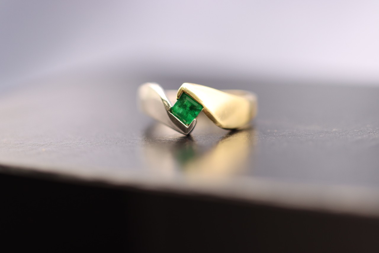 An emerald solitaire engagement ring sits on a black table.